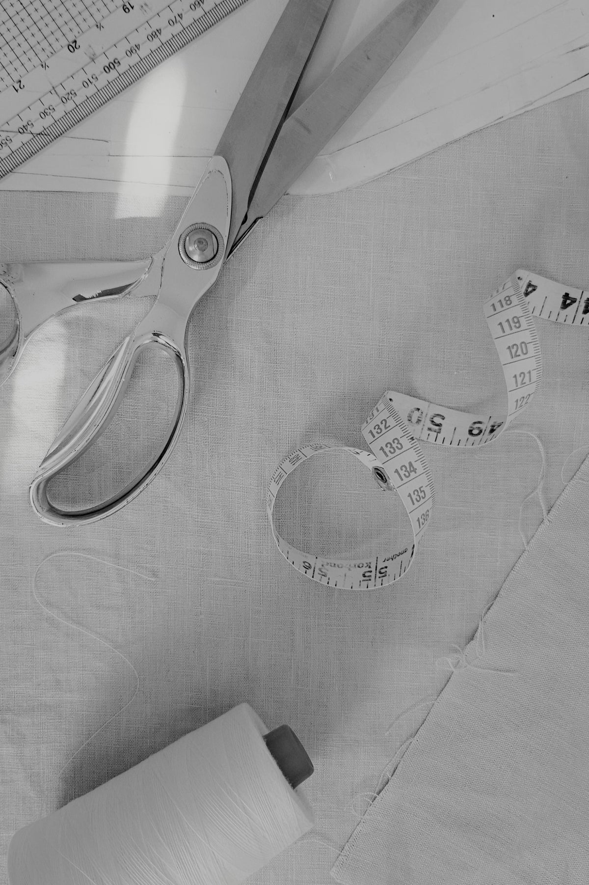 A black and white close-up of sewing and tailoring tools on fabric, including scissors, a measuring tape, a ruler, and a spool of thread.