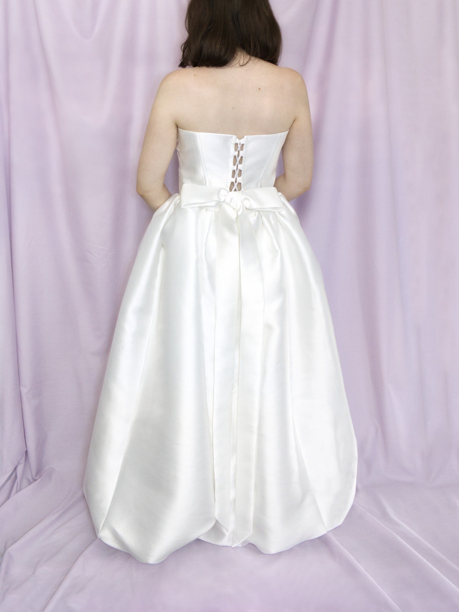 back view of a white lustrous strapless dress with lace up back on a model against a purple background.