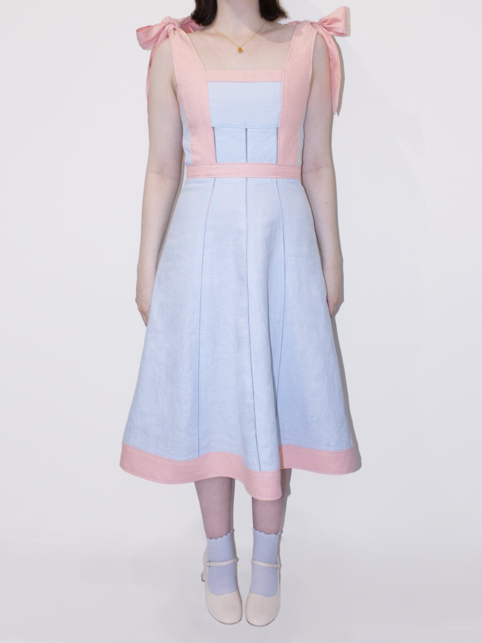 A linen A-Line midi dress with square neckline. The straps from the waistline, the waistband, and along the hem and neckline are light pink, the remaining is light blue. The straps are tied into large bows at the shoulders and there are three vertical rows of open stitching in the bodice and skirt.