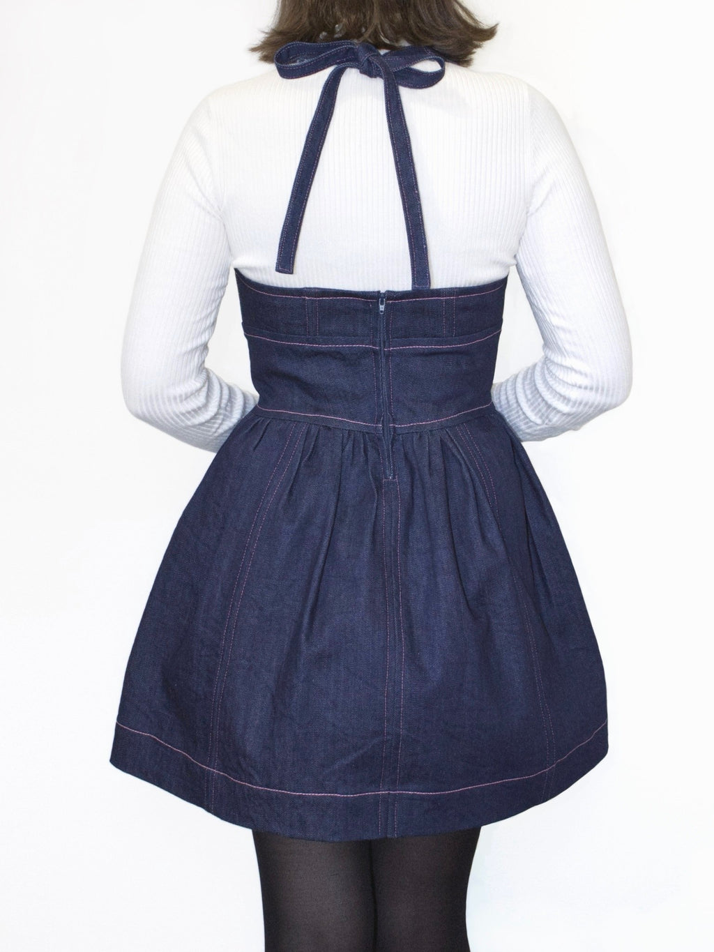 Back view of a denim mini dress showing the halter straps tied into a bow at the back of the neck. The back neckline goes straight across at mid back level and there is a centre back zip.