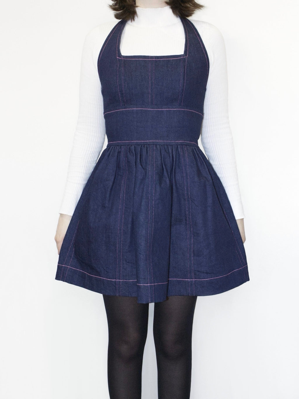 A dark wash denim mini dress with light pink topstitching along the neckline, armholes, waistline, hem and vertically along the seams. It has a square halter neckline and gathered A-line skirt. It is worn over a white long sleeve top and black tights.