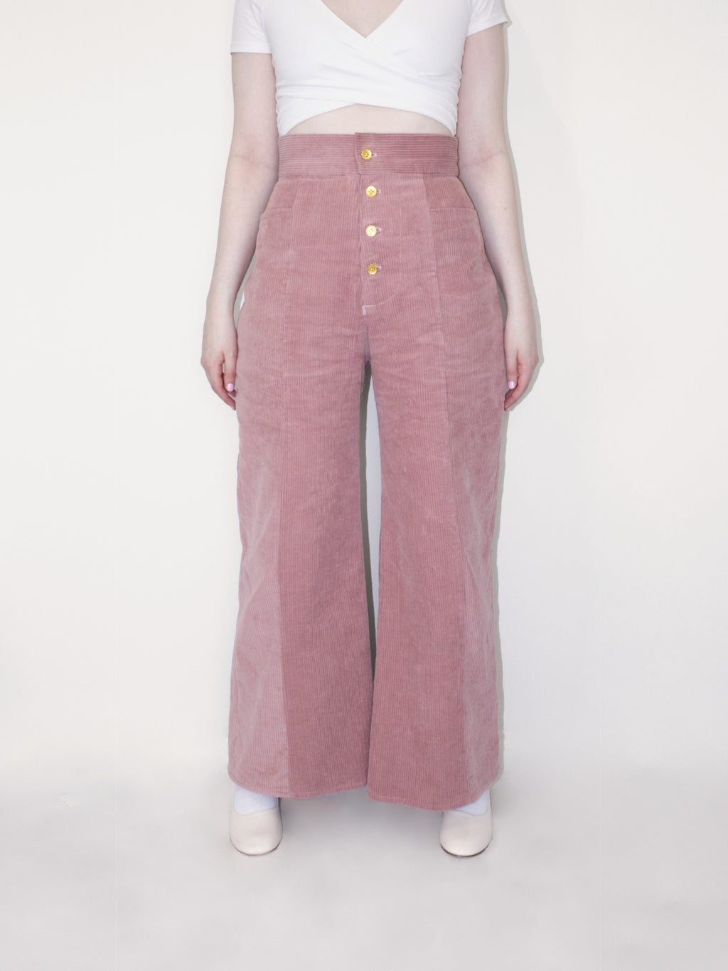 Pink corduroy pants with a high waisted and wide leg fit with seams down the centre of each leg. There are four gold buttons along the fly and two front pockets.