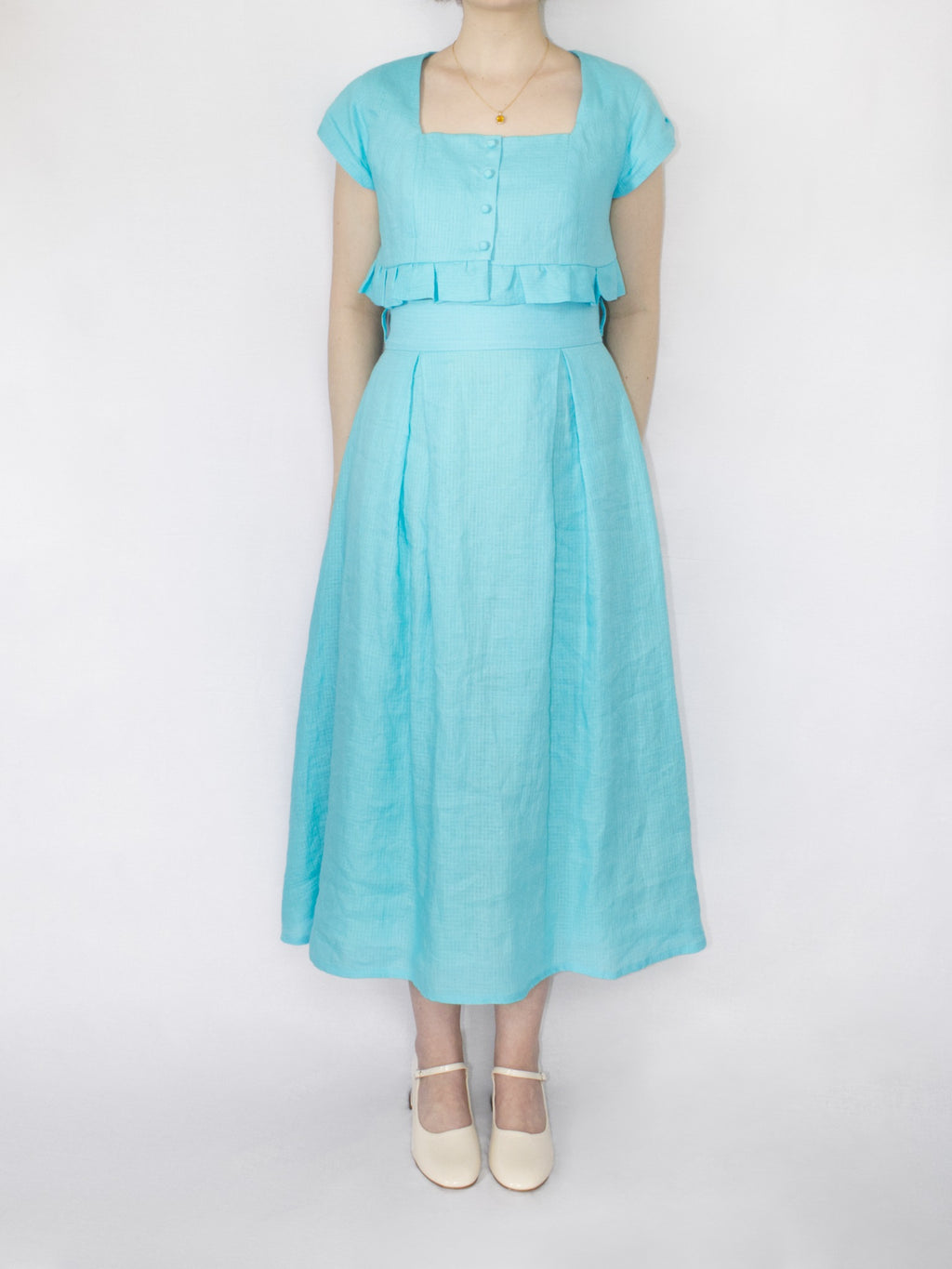 An aqua blue linen set consisting of a cropped button up top with frill hem and square neckline and a midi length high waisted skirt with a wide waistband and two box pleats on the front and back.