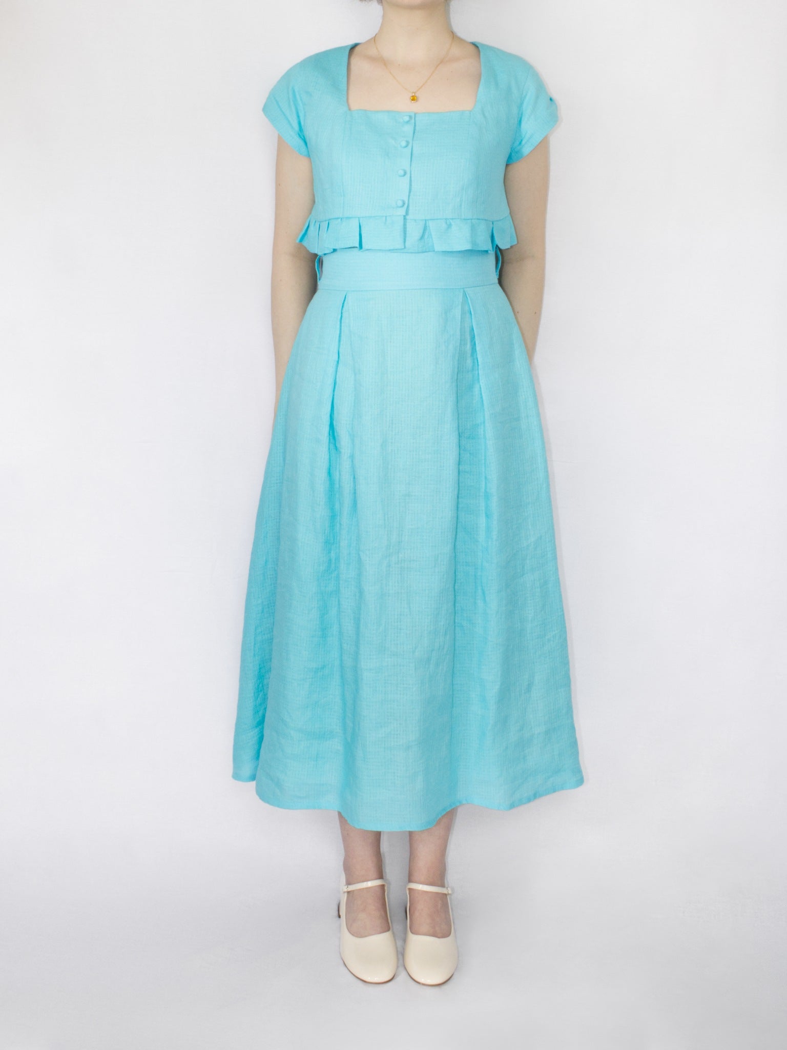 An aqua blue linen set consisting of a cropped button up top with frill hem and square neckline and a midi length high waisted skirt with a wide waistband and two box pleats on the front and back.