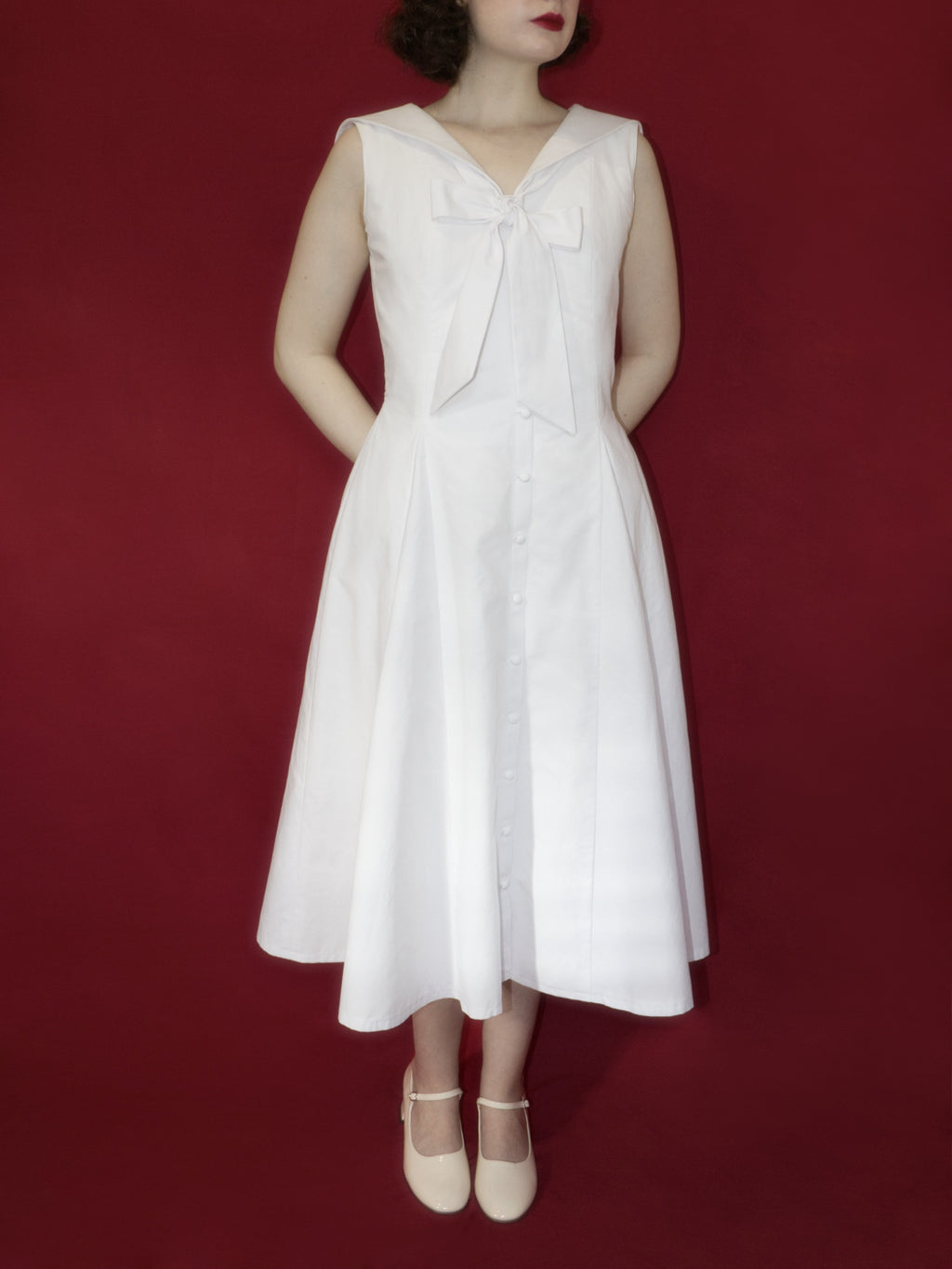 Front view of a sleeveless white button up midi dress with V-neckline, sailor collar, neck tie tied into a large bow, and full A-Line skirt.
