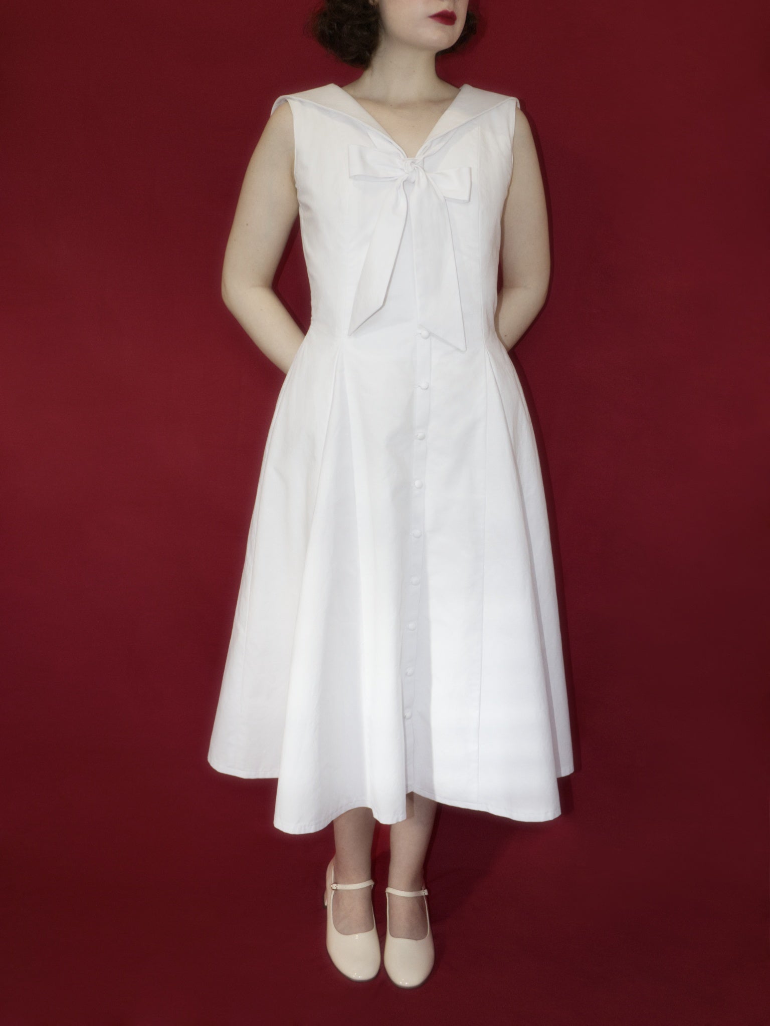 Front view of a sleeveless white button up midi dress with V-neckline, sailor collar, neck tie tied into a large bow, and full A-Line skirt.