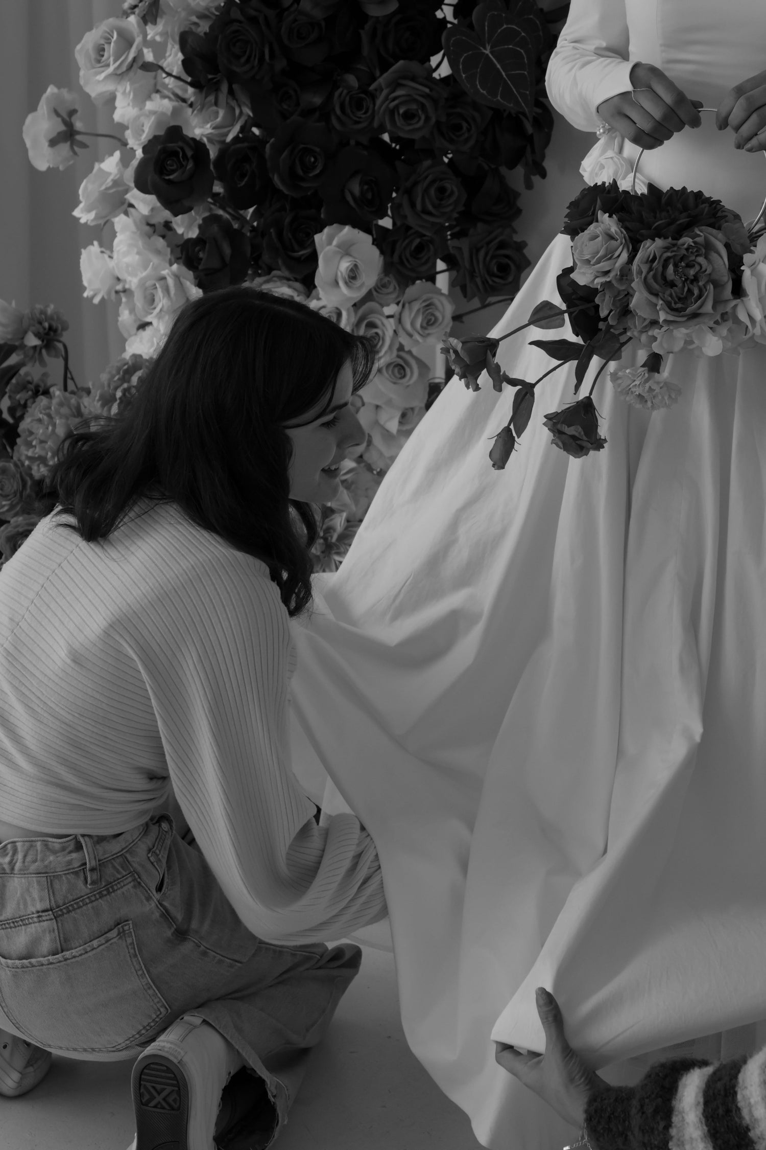 Black and white photo of a woman kneeling to adjust the skirt of a white wedding dress, with a large floral backdrop behind them.