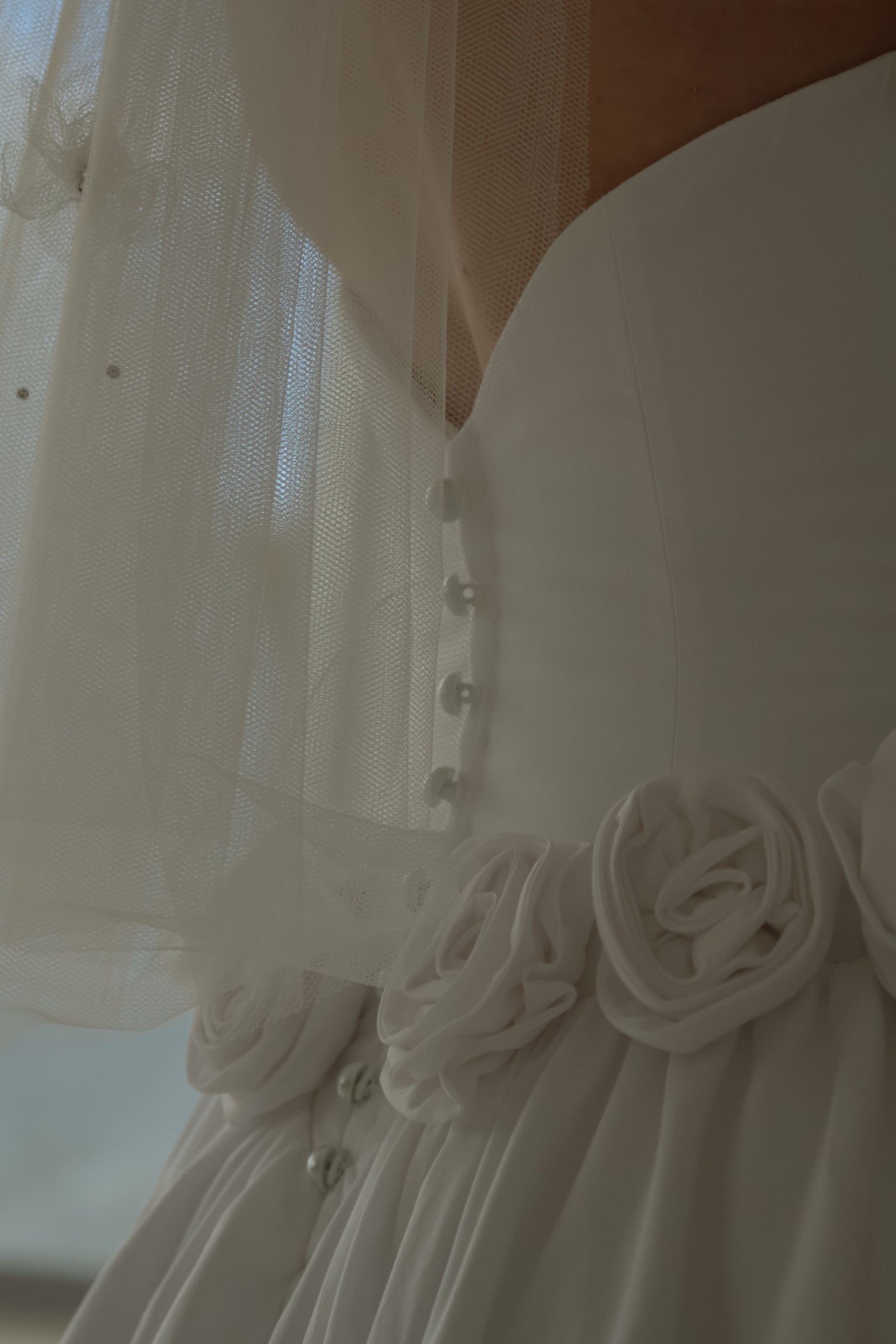 Close-up of a white bridal dress with a sheer tulle veil, pearl buttons, and fabric roses at the waist.