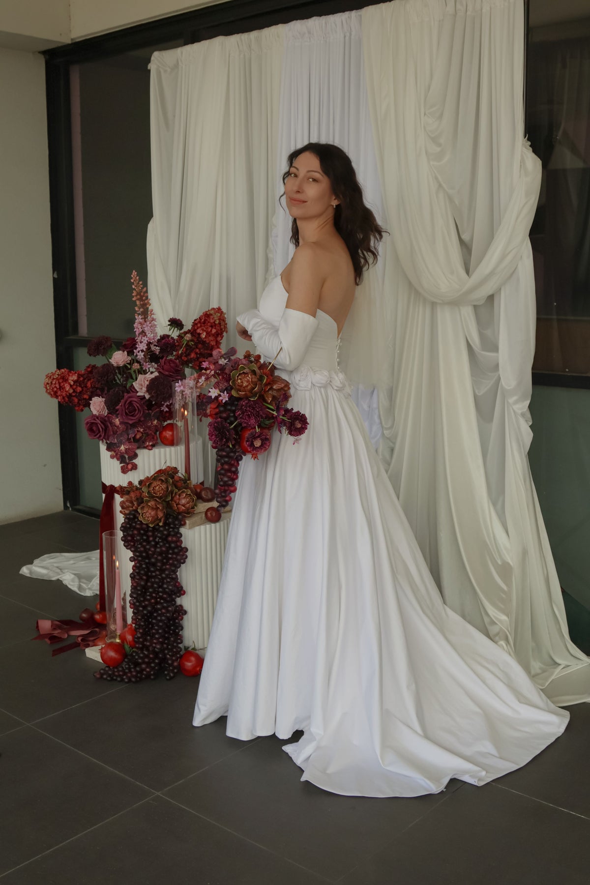 A woman wearing a strapless white wedding gown with long sleeves and a full skirt stands indoors in front of draped white fabric. She is holding a large floral arrangement with deep red, burgundy, and brown flowers and dark grapevine details.