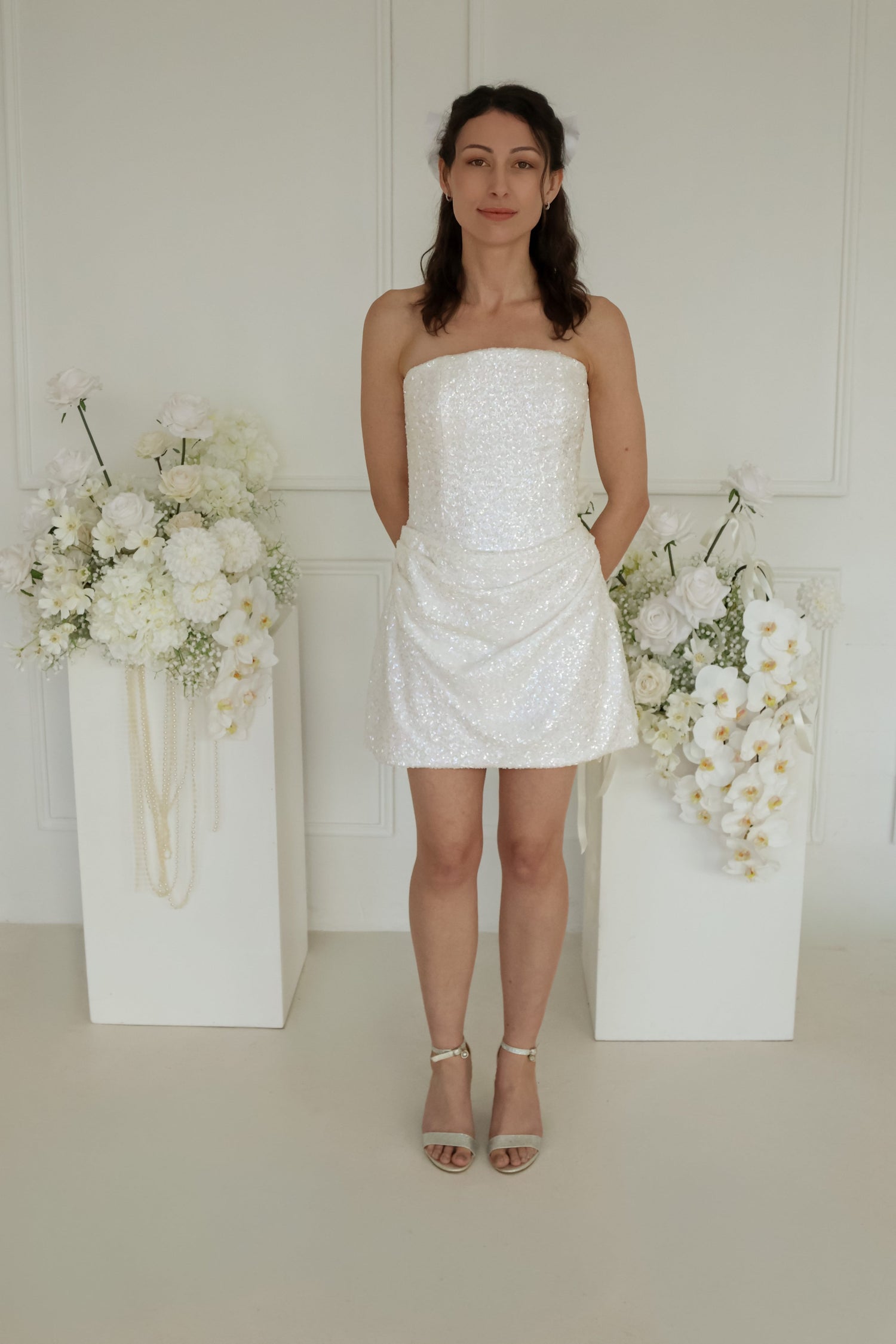 Full view of a woman wearing the moonlight mini dress. She is centered between two white pedestals decorated with large white floral arrangements against a white paneled wall.