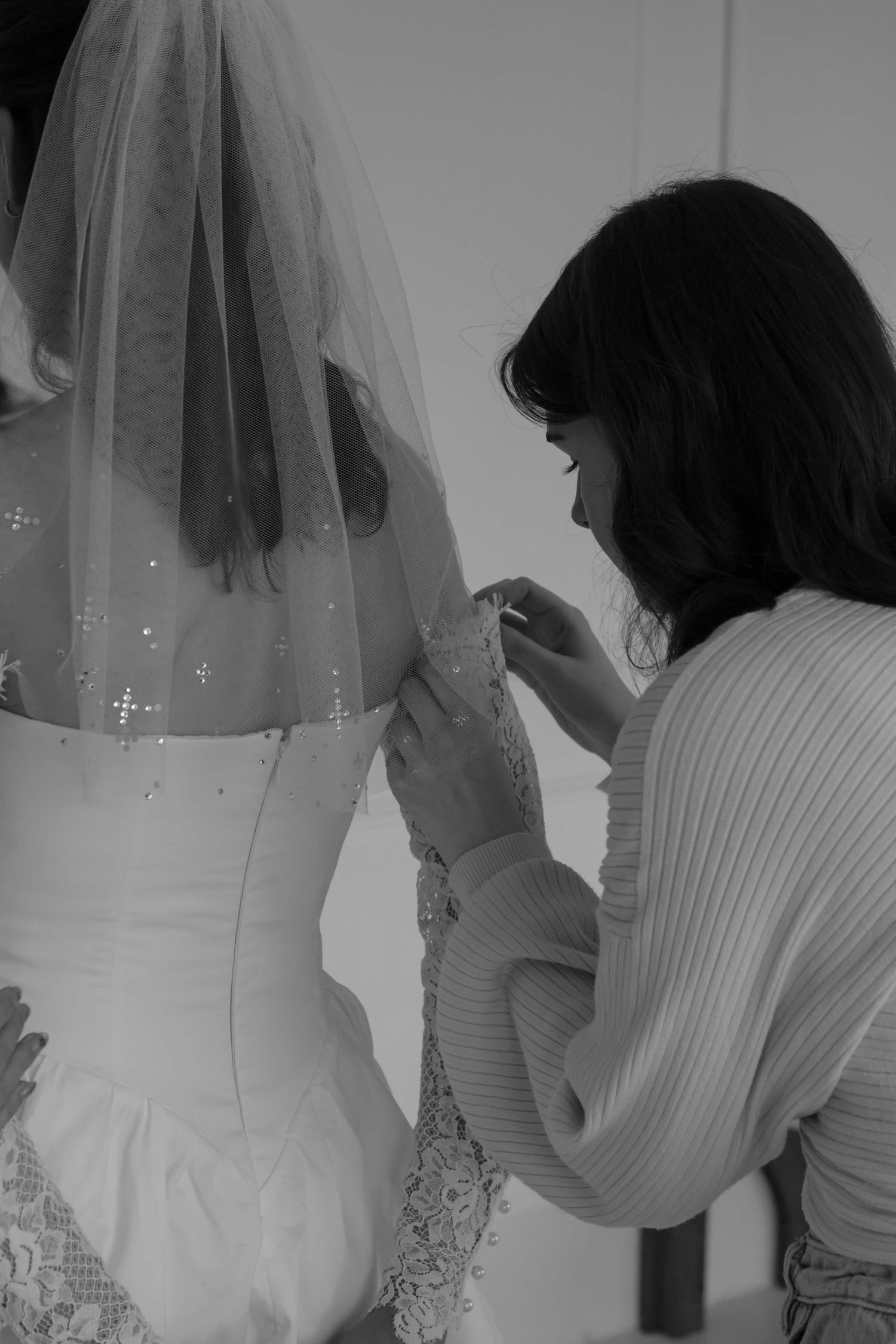 Black and white photo of a bride in a wedding dress and veil having her dress adjusted by a woman.