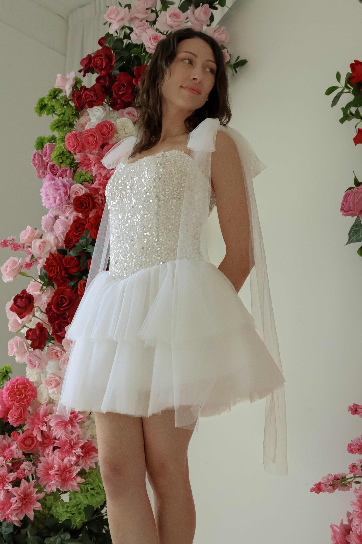 A woman wearing the starlight mini dress posing in front of a pink and red floral arch backdrop shot from a low angle, highlighting the long length of the tie-able tulle straps as they cascade down below the skirt.