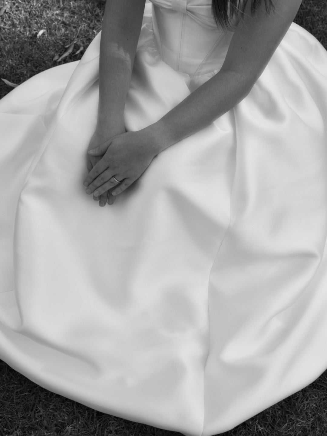 A black and white close-up of a woman seated on grass, wearing a voluminous mikado wedding gown with hands folded over the skirt showing her wedding ring.