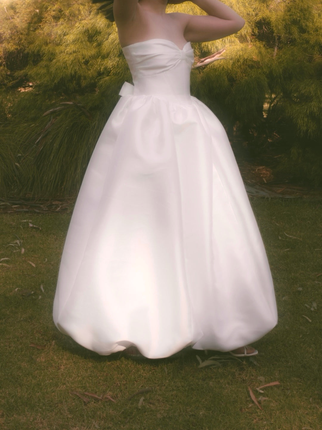 A woman in a grassy park wearing a strapless mikado bridal gown with gathered bow design at the bust and gathered floor length skirt with bubble hem.