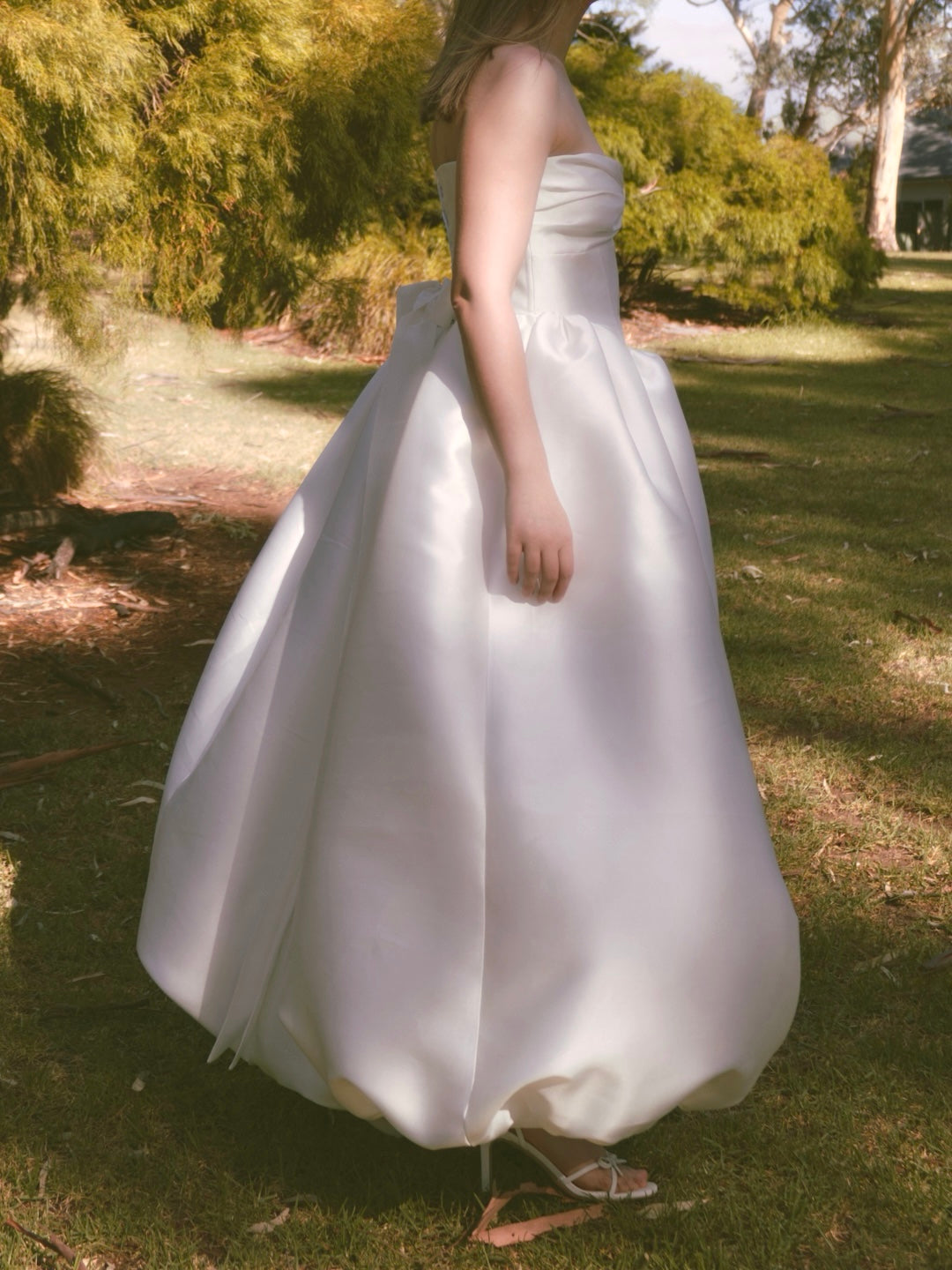 Side view of a woman outdoors in a grassy park wearing a strapless white mikado bridal gown with gathered bow design at the bust, a gathered floor length bubble hem skirt, and a large bow at the back waistline.
