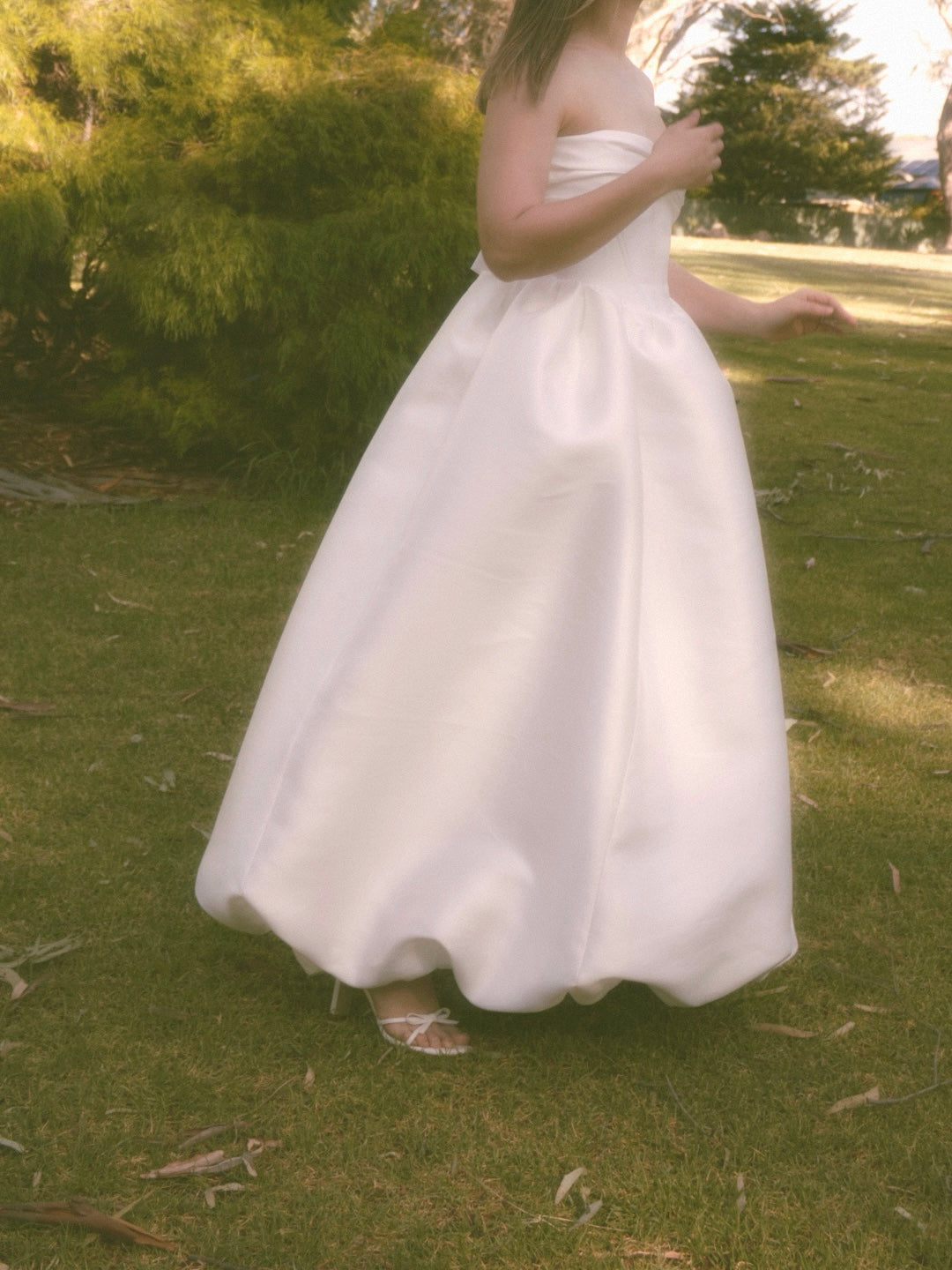 A woman outdoors on grass wearing a strapless white mikado bridal gown with gathered bow design at the bust and gathered floor length bubble hem skirt.