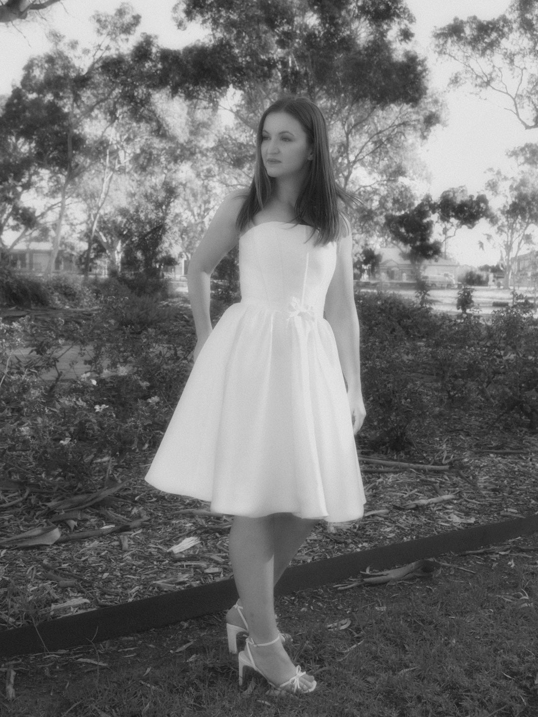 A woman wearing a white strapless square neckline dress with a fitted bodice and flared skirt with bow detail on the waistband, standing outdoors in a park-like setting with trees and a flower bed in the background. The photo is in black and white.