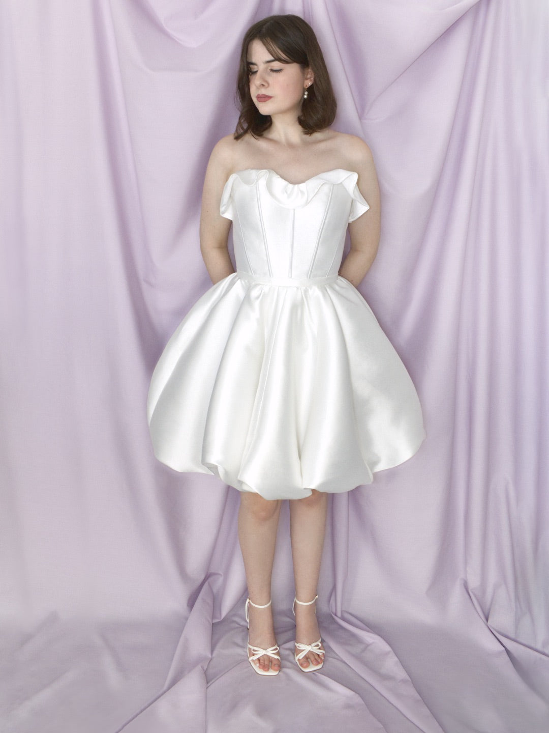 A woman modeling a strapless white mikado mini dress with a ruffled sweetheart neckline, a fitted waist, and a full flared bubble hem skirt, posed against a draped lavender fabric backdrop.