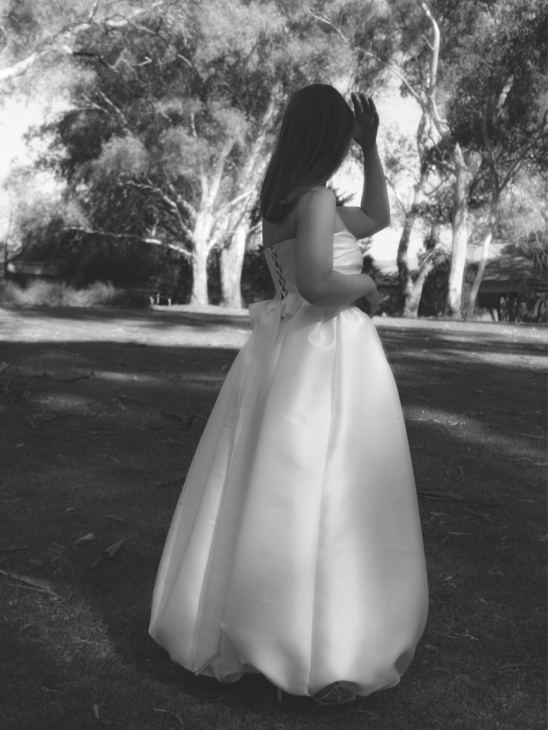 A woman wearing a strapless white mikado bridal gown stands outdoors in a park with trees in the background, photographed in black and white. The gown is floorlength with a bubble hem and a lace up back with a large bow at the back waistline.