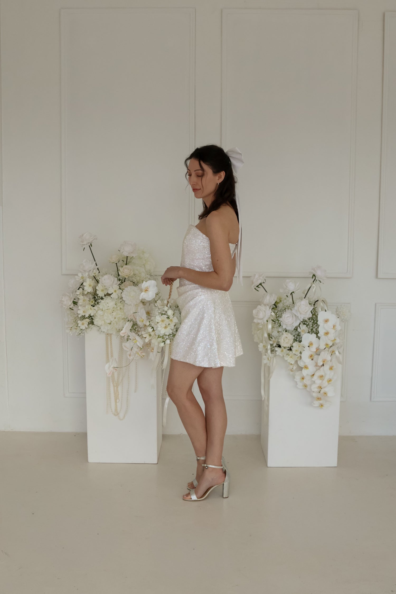 Side view of a woman wearing a strapless sequin mini dress and large white hair bow in front of white floral arrangements on pedestals.