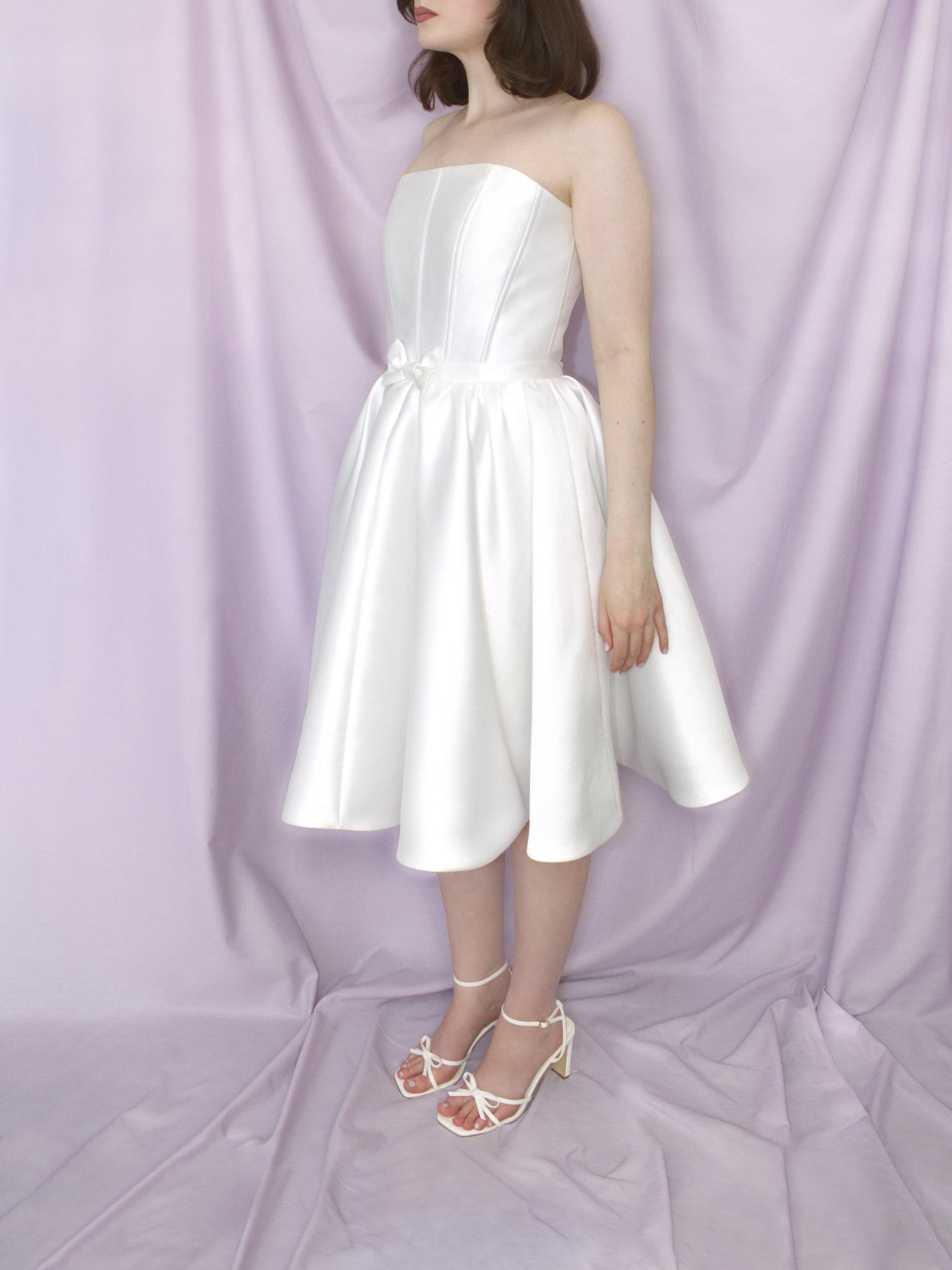 Woman wearing a white mikado mini dress with square neckline, fitted waist, flared circle skirt and bow detail on the waistband modeled against a draped lilac fabric backdrop.